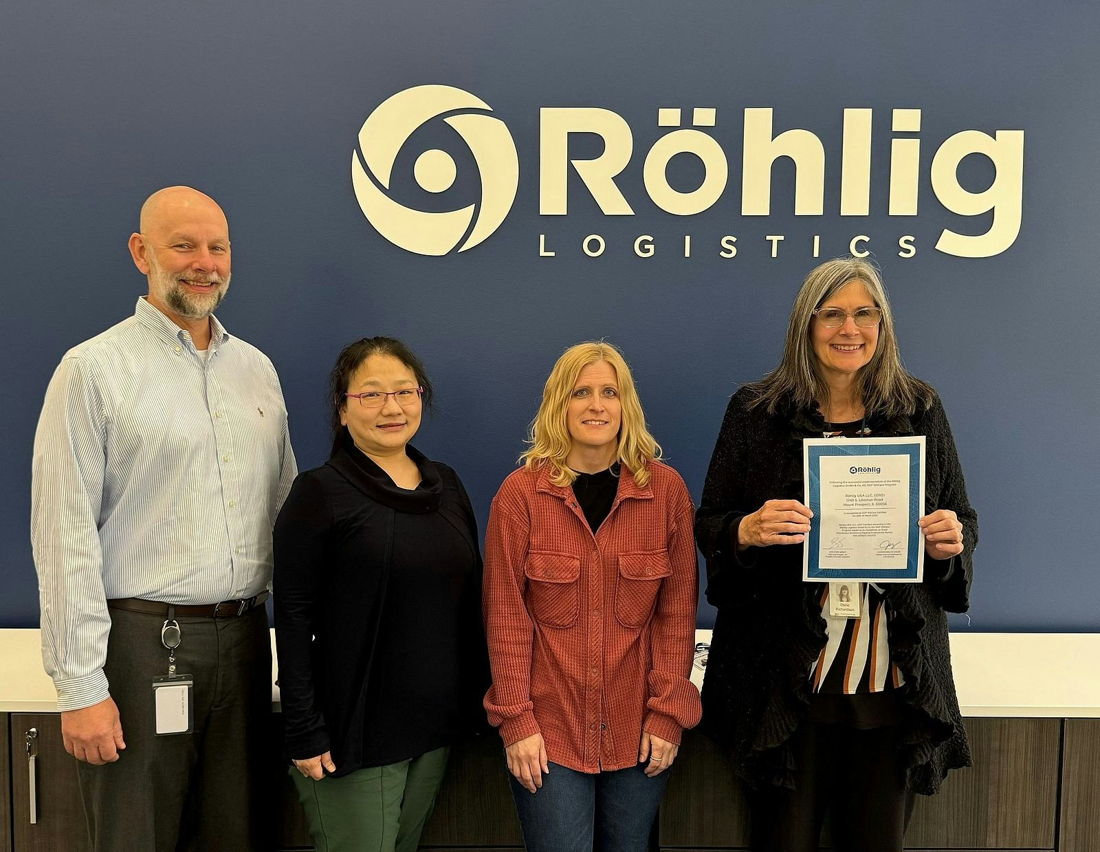 Vier Personen stehen vor einer blauen Wand mit dem Röhlig-Logo. Eine von ihnen hält ein Zertifikat in der Hand.
