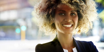 A woman with blonde curly hair, brown eyes and a nose piercing is wearing a black blazer and white blouse. She is smiling in the camera.
