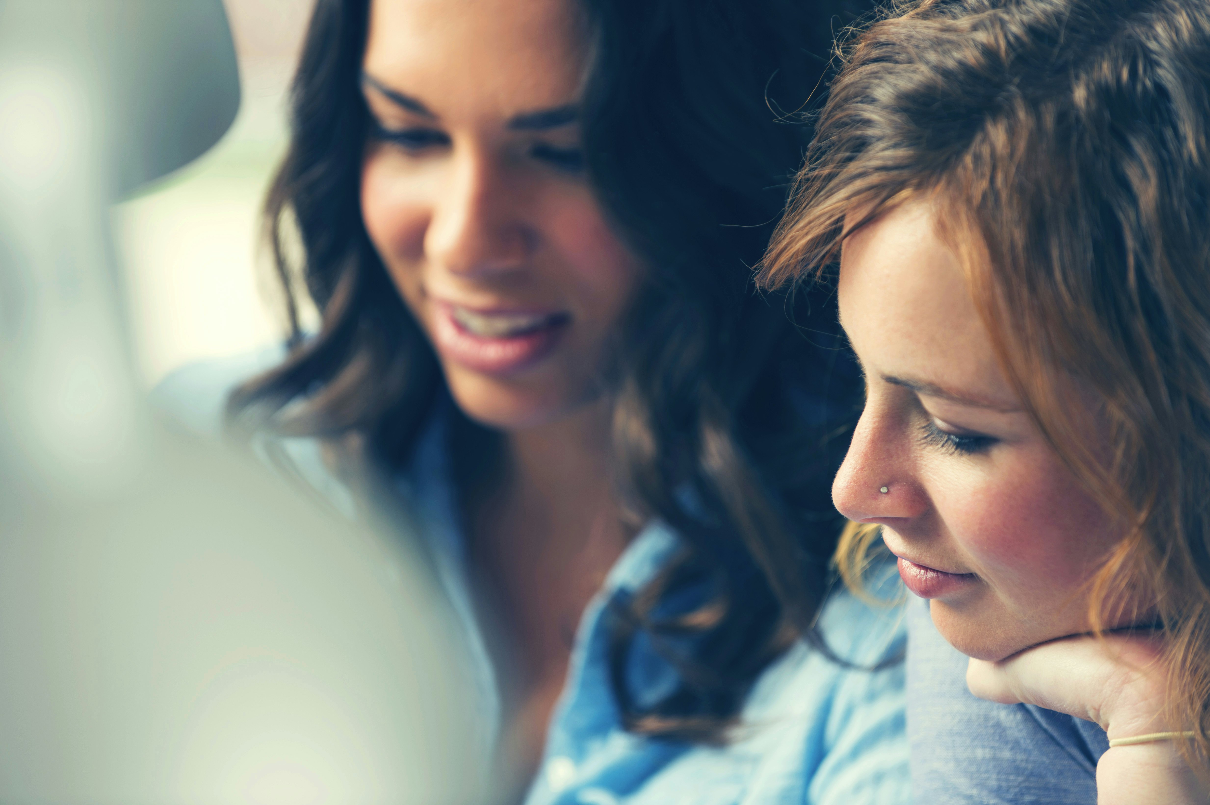 Two women with brown hair and blue shirts looking on something. One has a nosepircing.