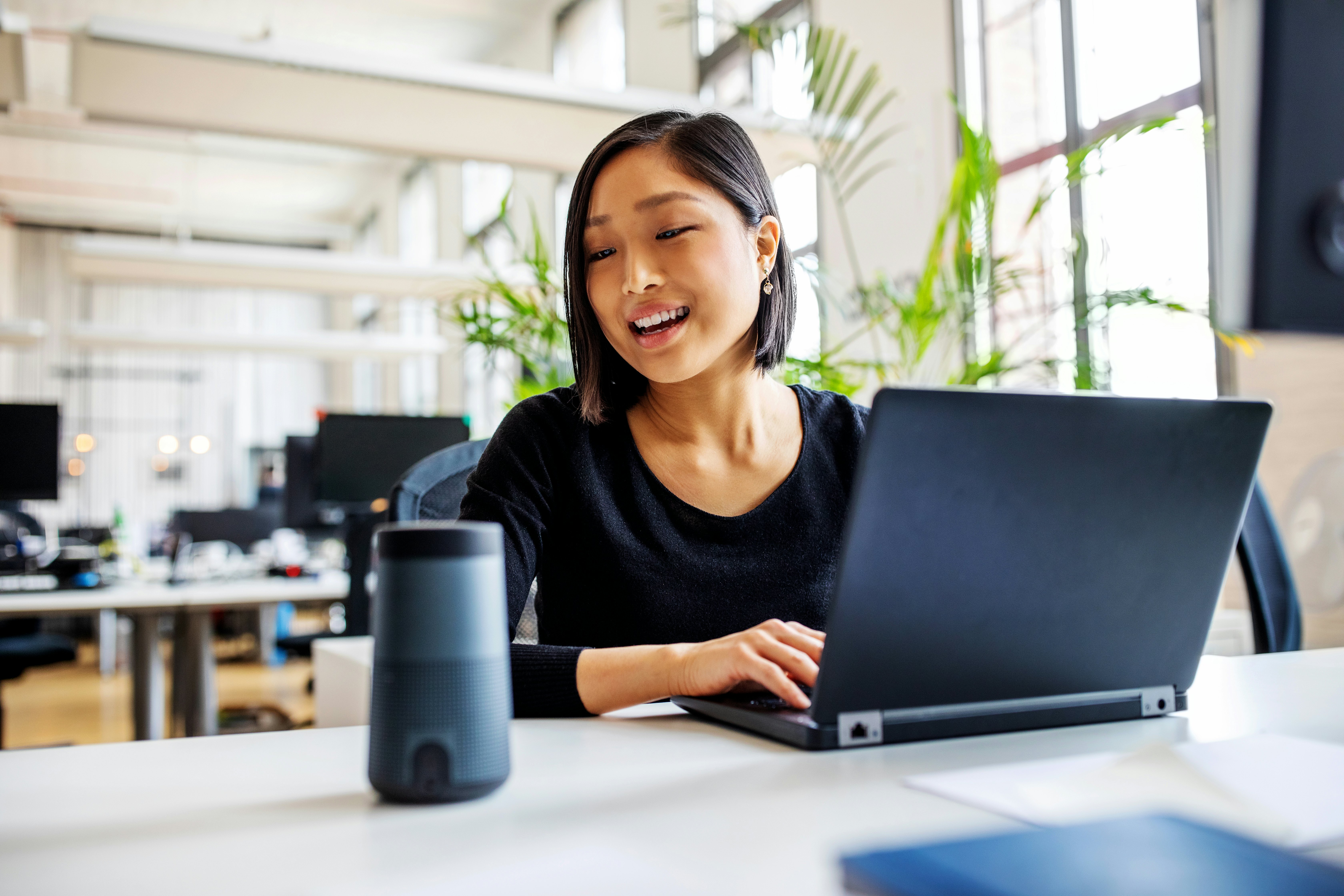 A woman in an office with brown hair wearing a black shirt is sitting at a desk in front of a black laptop looking at a black speaker.