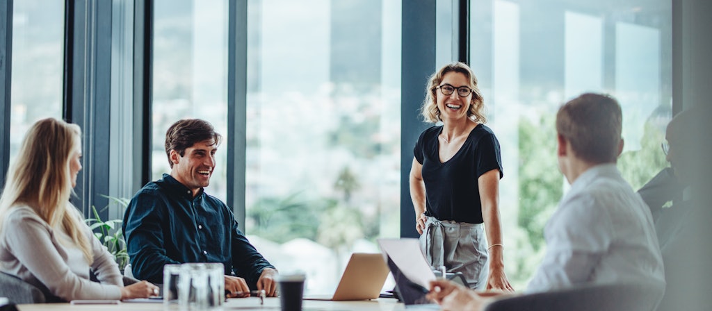 Four people are at an office Desk and one Women with Glasses is standing right next to them. All are laughing.