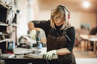A Woman with hearing protection and safety glasses wearing a work apron and gloves is drilling into a piece of wood with a cordless drill.
