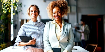 Two smiling women are standing in a room in front of a table. One has brown curly hair wearing a lightblue jacket holding a notebook and the other one has blonde hair wearing a blue and white stripped shirt and rose pants holding a tablet.