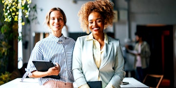 Two smiling women are standing in a room in front of a table. One has brown curly hair wearing a lightblue jacket holding a notebook and the other one has blonde hair wearing a blue and white stripped shirt and rose pants holding a tablet.