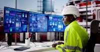 A worker with a constructions helmet and neon yellow jacket sits at a Desk in front of three monitor screens showing some systems.
